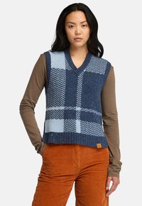Gebreide mouwloze vest in donkerblauw met lichtblauwe ruitpatronen, met een V-hals en ribbelrand, gecombineerd met een lange mouw bruin shirt.