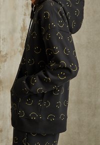 Black hoodie made of cotton blend, featuring yellow smiley face pattern. Includes a front pocket and a drawstring hood.