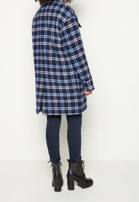 Mörkblå rutig kappa med ljusblå och vita accenter, enkelknäppt, ihop med mörka jeans och tjocka svarta klackstövlar.