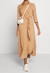 Abito lungo con bottoni in tessuto marrone chiaro con righe verticali bianche, caratterizzato da una vita annodata e maniche a tre quarti, abbinato a sneaker bianche.