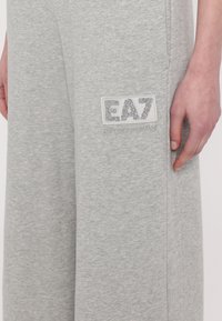 Grijze katoenen sweatpants hebben een brede pijp ontwerp. Een gestikte EA7-logo in zilver glitter is prominent weergegeven aan de linkerkant.