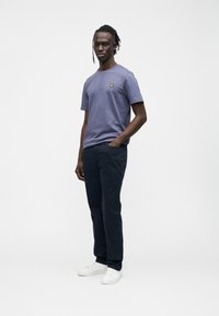 Camiseta de algodón de manga corta en color púrpura claro con un logo amarillo en el pecho, combinada con pantalones azul marino oscuro y zapatillas blancas.