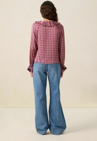 Frau mit welligem braunen Haar steht mit dem Rücken zur Kamera, trägt eine langärmlige lila gemusterte Bluse und hoch taillierte, weite blaue Jeans.