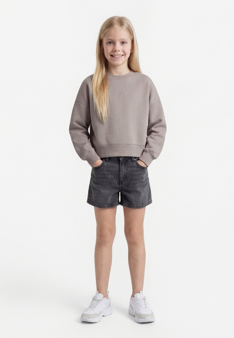 Egy szőke lány áll mosolyogva, kezét zsebre dugva, szürke pulóvert, fekete farmer rövidnadrágot és fehér tornacipőt visel tiszta fehér háttér előtt.