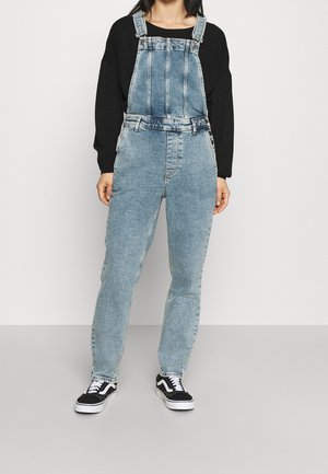 Henkilö, joka on pukeutunut vaaleansinisiin farkkuhaalareihin, mustaan pitkähihaiseen paitaan ja musta-valkoisiin tennareihin, seisoo vaaleaa taustaa vasten.