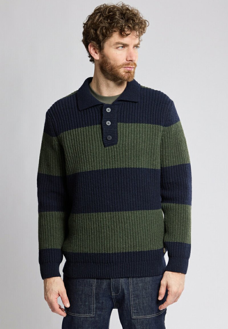 Homme avec des cheveux bouclés et une barbe, portant un pull côtelé à boutons avec de larges rayures horizontales bleu marine et vert, associé à un jean en denim noir.