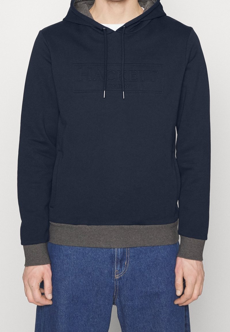 Homme portant un sweat à capuche bleu marine avec des poignets et un ourlet gris, des cordons bleu marine et un jean bleu, debout les bras détendus le long du corps.