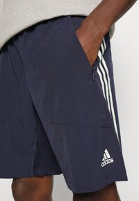 Marina atletiska shorts med ljusgrå tre-stripes design. Har dragkedjorfickor och en Adidas-logga i nederkanten. Slät textur.