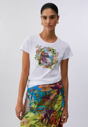Donna con una t-shirt bianca con stampa di una scena di spiaggia colorata e una corona floreale, abbinata a una gonna floreale multicolore vivace.