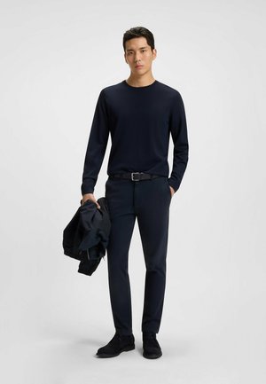 Chemise à manches longues marine, pantalon marine et chaussures foncées. Le modèle tient une veste foncée. Tissu lisse, coupe ajustée et design minimaliste.
