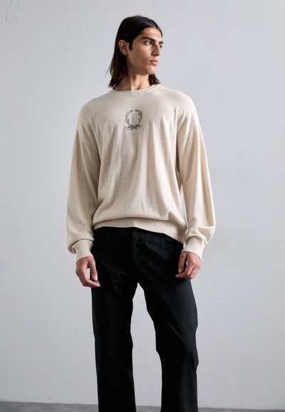Suéter de punto beige con mangas largas y puños acanalados, que presenta un emblema circular negro bordado en el pecho, combinado con pantalones negros.