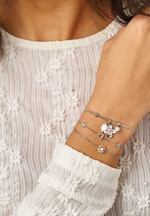 Handgelenk einer Frau, die drei zarte silberne Charms-Armbänder mit Herzen, Blumen, Perlen und rosa Edelsteinen trägt, vor einem weiß strukturierten Strickärmel.