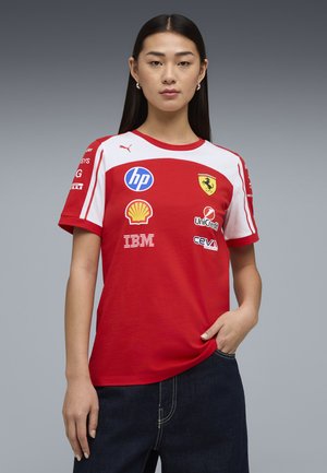 Ung kvinde med langt mørkt hår iført en rød og hvid Ferrari racing T-shirt med sponsorlogoer, stående foran en ensfarvet grå baggrund.