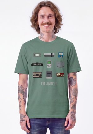 Homme avec des tatouages et des piercings au nez portant un t-shirt vert avec des consoles de jeux rétro et le texte "I'm lovin' it".