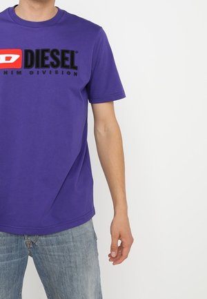 Hombre con camiseta morada de Diesel con texto de logo en negro y rojo, combinado con vaqueros azul claro, sobre un fondo blanco.