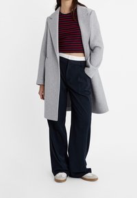 Lichtgrijze overjas, cropped top met rode en marineblauwe strepen, donkere wijde broek, en grijze sneakers met witte veters en beige accenten.