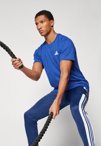 Blue athletic t-shirt with crew neck and Adidas logo, paired with blue pants featuring white stripes. Individual holding a black training rope.