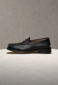 Μαύρος δερμάτινος penny loafer με ραφές μοκασίνου, χαμηλό τακούνι και ριγωτή εσωτερική επένδυση, εκτεθειμένος σε ουδέτερη επιφάνεια.