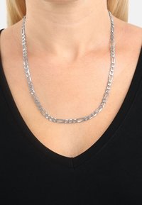 Collier en chaîne argentée avec des maillons allongés et ovales porté par une personne vêtue d'un haut noir à col en V.