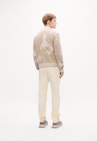 Young man standing, wearing beige and white patterned sweater, cream pants, and light sneakers, facing sideways on a white background.