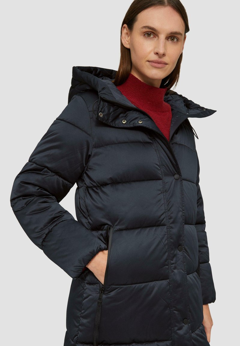 Marineblaue Pufferjacke mit Kapuze, mit abgesteppten Einsätzen, einem Reißverschluss vorne und Seitentaschen. Der Stoff wirkt glatt und isoliert.