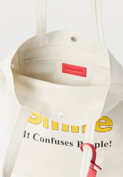 Κρεμ καμβά τσάντα ώμου με το κείμενο "Χαμογέλα, μπερδεύει τους ανθρώπους!" σε κίτρινο και μαύρο, κόκκινη ετικέτα επωνυμίας και λεπτομέρεια κόκκινου λουριού.