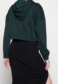 Sudadera de color verde oscuro, recortada, con capucha texturizada, mangas largas y puños acanalados, combinada con una falda negra acanalada con una abertura.