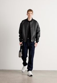 Chaqueta bomber de cuero negro con cremallera frontal, puños acanalados y un parche en la manga; combinada con jeans oscuros y zapatillas blancas.