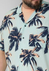 Korte mouwen shirt in lichtblauw met zwart en perzik palmboomprint, voorzien van een borstzak en zwarte knopen.