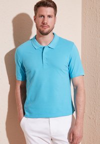 Camiseta polo azul brillante de manga corta, con cuello clásico y tapeta de tres botones. Llevada con pantalones blancos sobre un fondo texturizado.