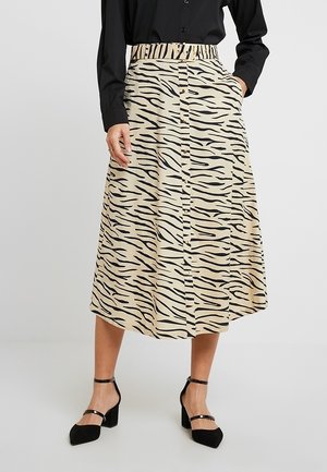 Jupe midi beige avec un motif rayé tigre noir, taille élastique et devant avec des boutons. Portée avec des talons noirs à brides de cheville.