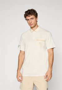 Beige poloskjorte laget av mykt stoff, med knapping på kragen, brystlomme med logo, og grønn brodering på ermet.
