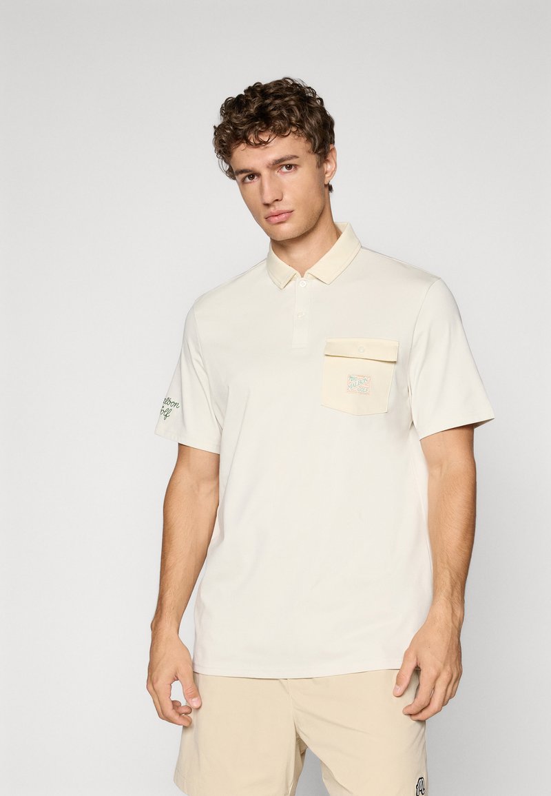 Beige poloskjorte laget av mykt stoff, med knapping på kragen, brystlomme med logo, og grønn brodering på ermet.