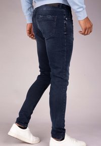 Jeans de mezclilla azul oscuro con un corte ajustado, que presentan un clásico diseño de cinco bolsillos y un ligero desgaste en el bolsillo trasero. Combinados con zapatillas blancas.