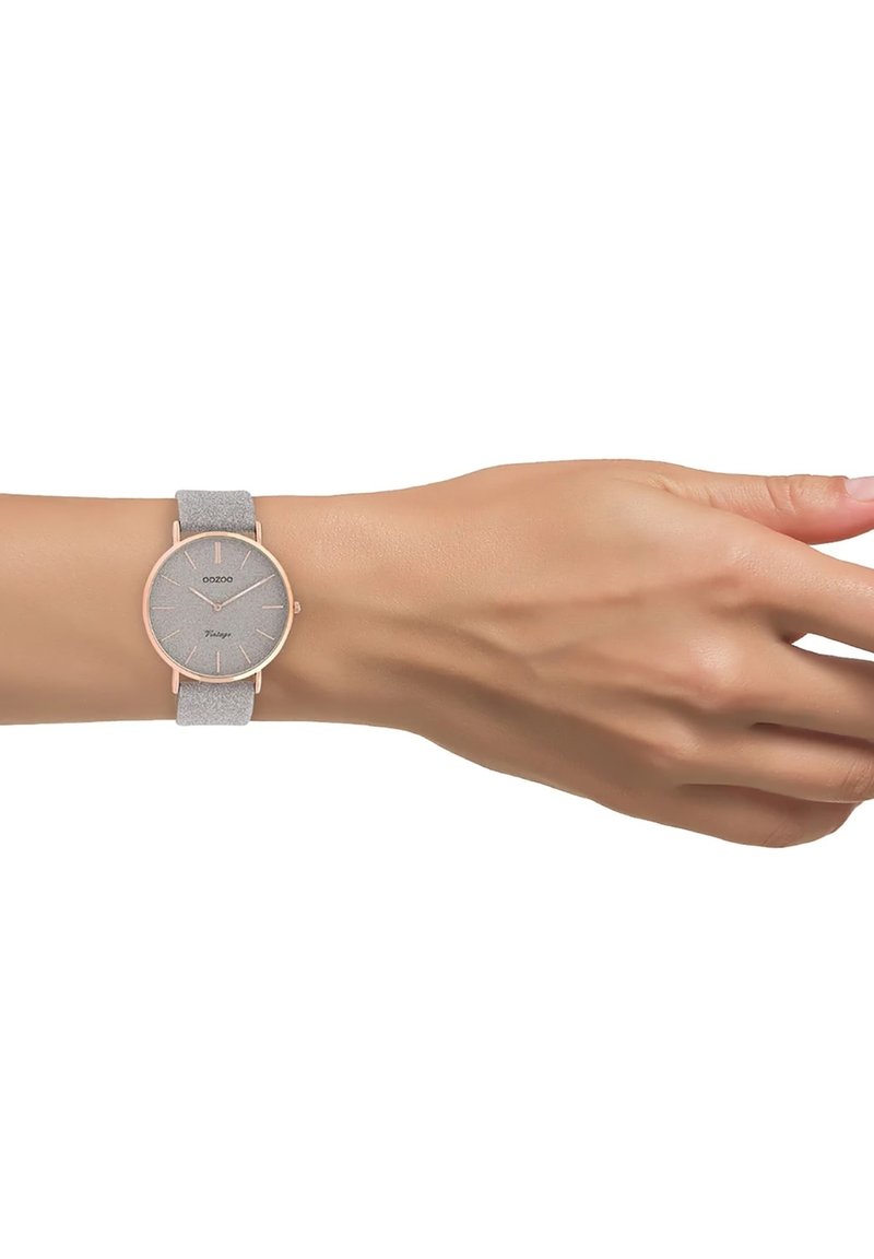 Montre analogique ronde avec un bracelet en tissu gris, des accents en métal rose gold et un cadran gris texturé avec des marqueurs d'heure minimalistes.