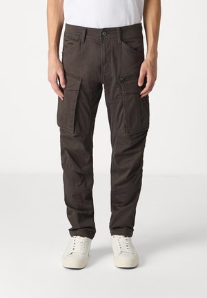 Cargo pants in dark gray fabric, featuring multiple side pockets, zipper details, a button closure, and tapered leg design. Paired with white sneakers.