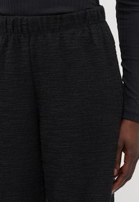 Black textured trousers with an elastic waistband, made of a soft material. The fabric has a slightly uneven surface pattern.