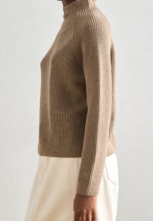 Personne portant un pull à col roulé côtelé marron moulant et une jupe beige clair sur un fond uni clair.