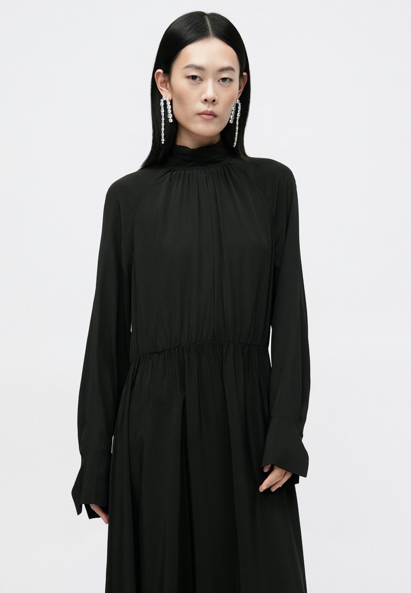 Vestido negro de manga larga con cuello alto, cintura fruncida y falda fluida. Textura suave con un ligero brillo. Presenta mangas acampanadas y drapeado.