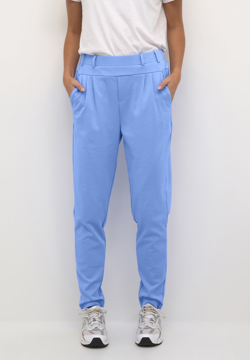 Pantaloni tapered de culoare albastră deschisă, confecționați dintr-un material moale, cu buzunare laterale, față plată și o textură netedă. Purtați cu adidași argintii.