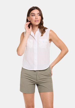 Mujer vestida con camisa blanca sin mangas abotonada con bolsillos delanteros y pantalones cortos de color verde oliva, de pie con una mano en la cadera y la otra cerca de su oreja.