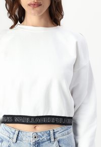 Witte cropped sweatshirt met lange mouwen en uitsparingen op de schouders. Voorzien van een zwarte elastische band met het logo "KARL LAGERFELD JEANS".