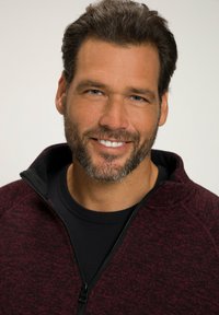 Giacca zip-up da uomo in tessuto rosso scuro e nero melangiato, con colletto alto e cerniera completa, indossata sopra una maglietta nera. Capelli corti e barba.