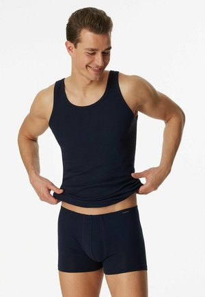 Mann passt marineblaues Tanktop und passende Boxershorts an, lächelt, vor weißem Hintergrund.