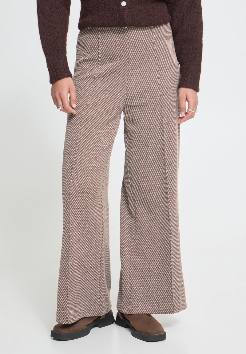 Personne portant un pantalon rayé marron taille haute à jambes larges, un pull marron foncé et des chaussures plates marron, debout sur un fond uni.