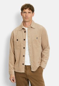 Beige Cordjacke mit Knopfverschluss, zwei Brusttaschen und Kentkragen. Getragen über einem weißen Hemd, kombiniert mit braunen Hosen.