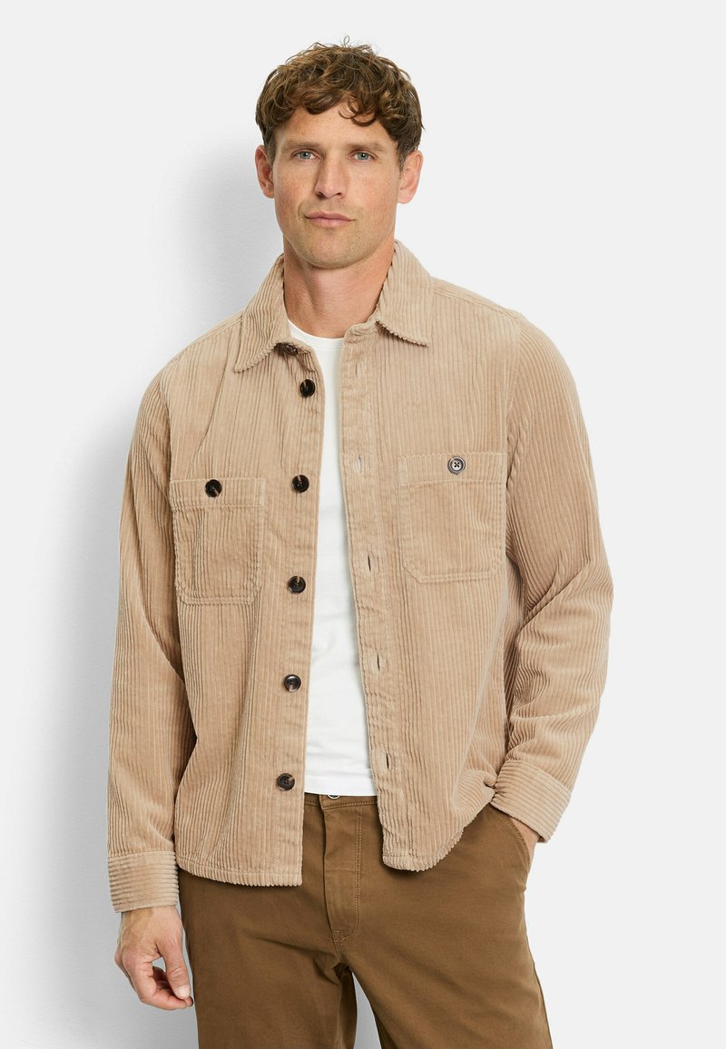 Beige Cordjacke mit Knopfverschluss, zwei Brusttaschen und Kentkragen. Getragen über einem weißen Hemd, kombiniert mit braunen Hosen.