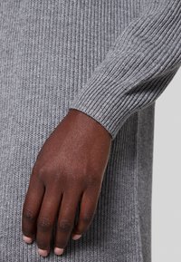 Tissu de pull gris côtelé, présentant un gros plan d'une texture tricotée et une main reposant sur la manche, mettant en valeur le détail du poignet.