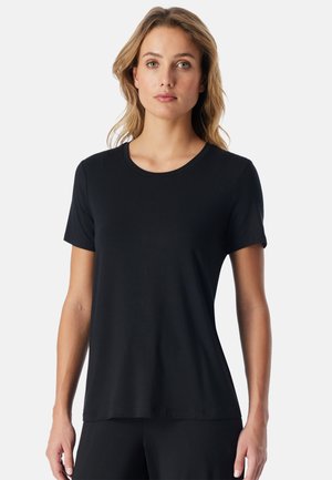 Vrouw met lichtblond haar die een eenvoudige zwarte korte mouwen T-shirt draagt, staat tegen een witte achtergrond en kijkt vooruit.