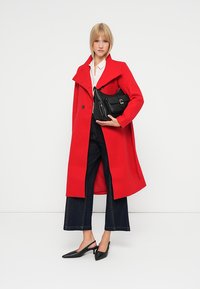 Cappotto rosso in lana con ampio colletto e bottoni, abbinato a una camicia bianca, jeans larghi scuri e scarpe slingback nere, mentre tiene una borsa nera.